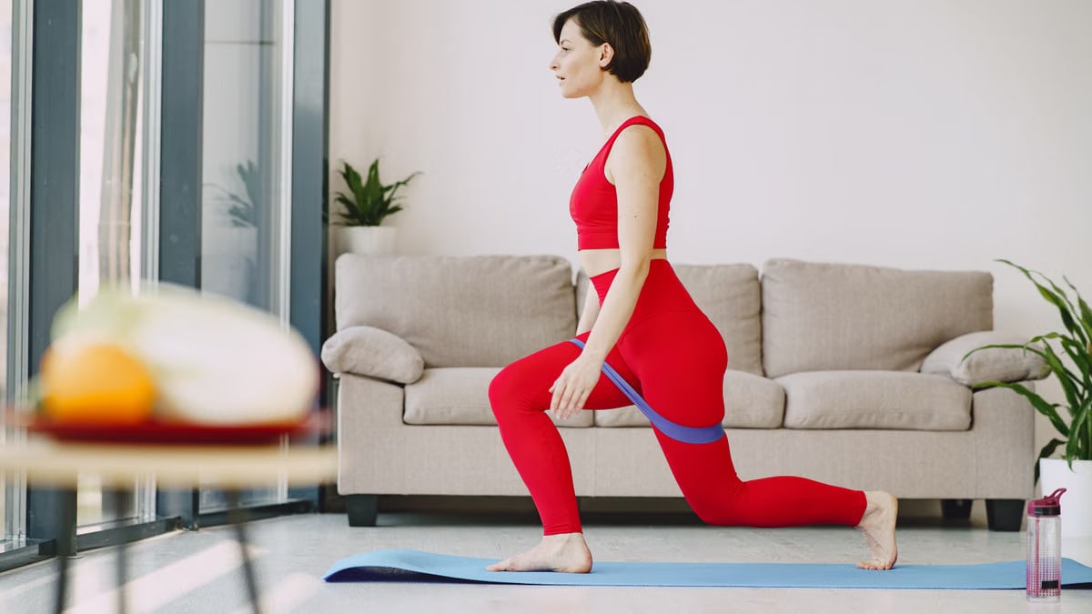 Side view of fit female in sportswear with short hair doing exercise with elastic band on blue mat near sofa in living room
